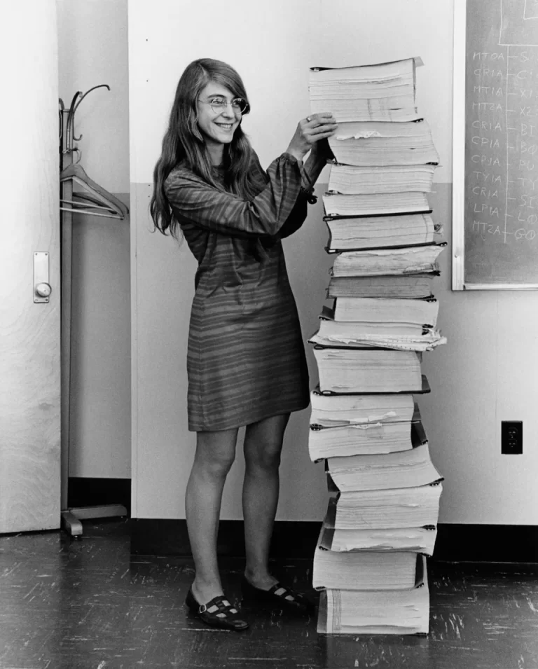 Black and white portrait of Margaret Hamilton smiling next to a stack of books